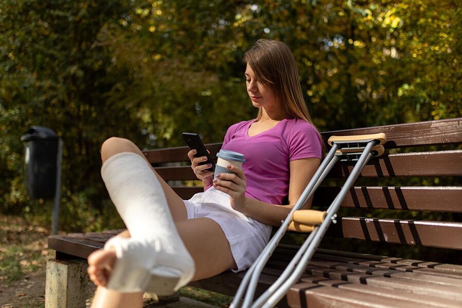 Woman with a leg cast and crutches sits on a bench holding a cup and phone.